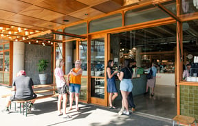 People hanging outside a cafe with large glass windows and doors.