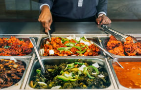 A staff member scooping up curry from a buffet behind glass.