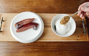 A hand spoons a garnish onto a dish on a wooden board.