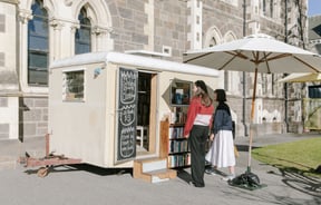 Two women standing outside the bookshop caravan on a sunny day.