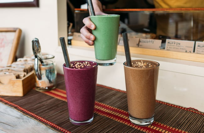A hand places a tall, berry-coloured smoothie on the counter beside glasses of a creamy green and a browny-peanut butter smoothie at Dripping Bowl.