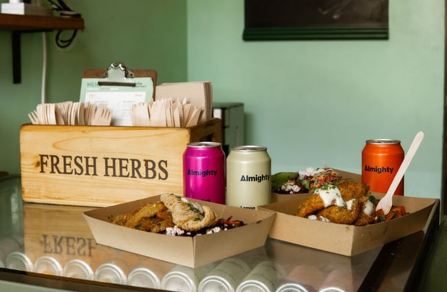Three meals and cans of drink on the counter at Herba Gourmet, with wooden takeaway utensils in a repurposed wooden crate in the background.