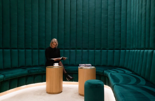 A blonde woman wearing black clothes sitting on a large green velvet seat.
