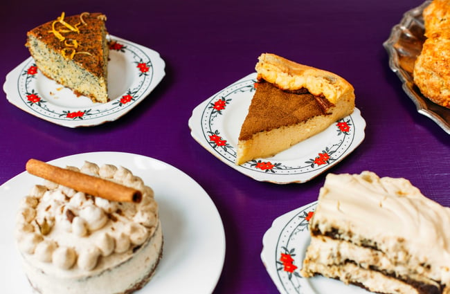 Different slices of cakes on china saucers on a table with a purple tablecloth.