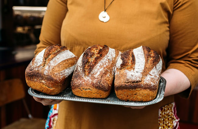 A lady holding a tray with three loaves of freshly baked bread.