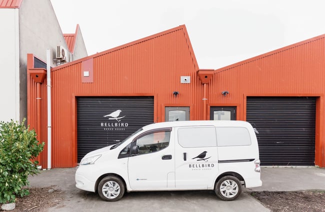 The back of the Bellbird Bakery premises, red corrugated walls ending in a saw tooth roof, with the white Bellbird delivery vehicle in the foreground.