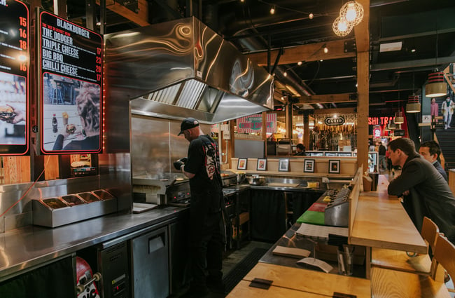 Chef in black shirt with logo standing over the grill cooking burgers.