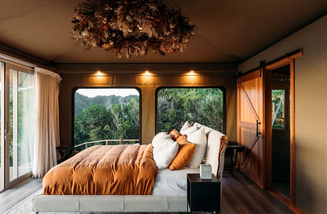 The bed and a small fireplace inside a little hut nestled into native bush.