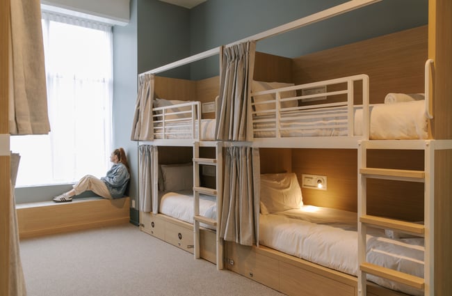 A woman sitting a room of bunk beds looking out a window.