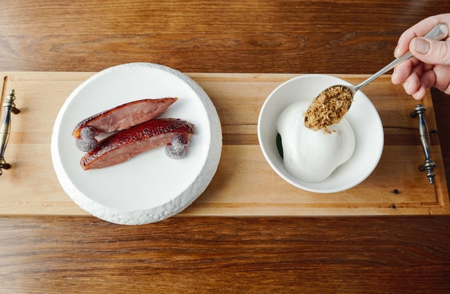 A hand spoons a garnish onto a dish on a wooden board.