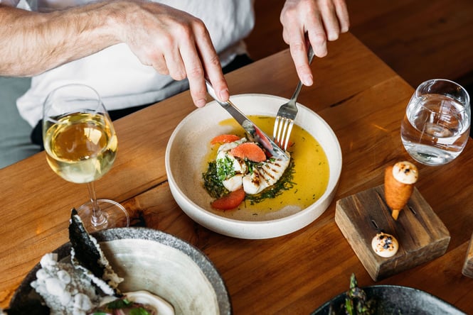 High angle view of a person cutting into a fish and citrus dish, wine and other dishes on the table at arc, Wānaka.