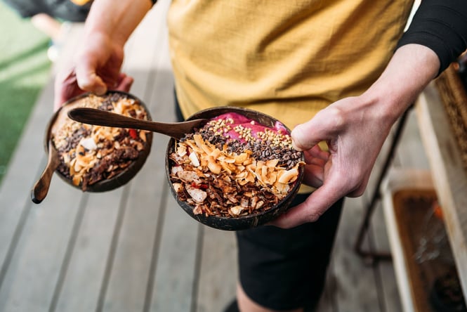 A staff member holds two bowls, made from coconut husks, of acai topped with crunchy coconut, cacao nibs and buckwheat groats.