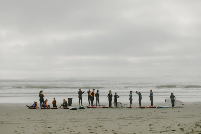 A group of kids standing along the shore with surfboards on a cloudy day.