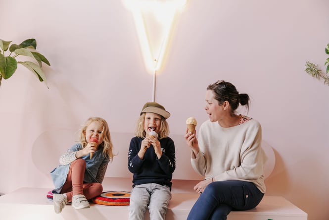 A woman and two kids eating ice cream in front of a pink wall.