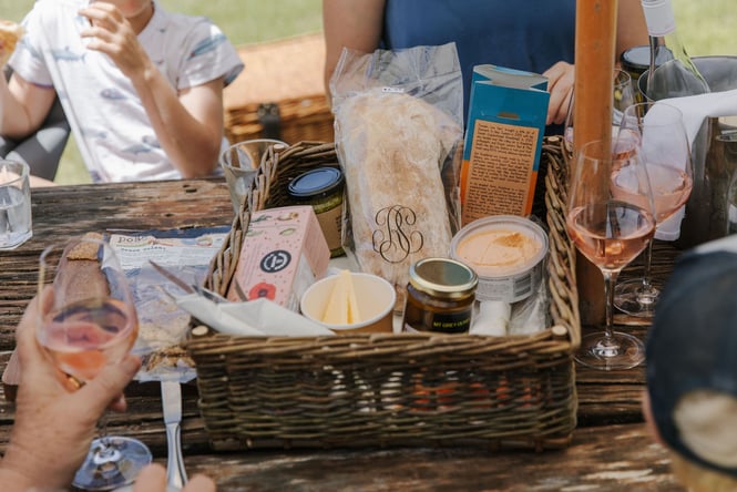 A close up of a basket of deli goods next to glasses of wine on an outside table at Pegasus Bay.