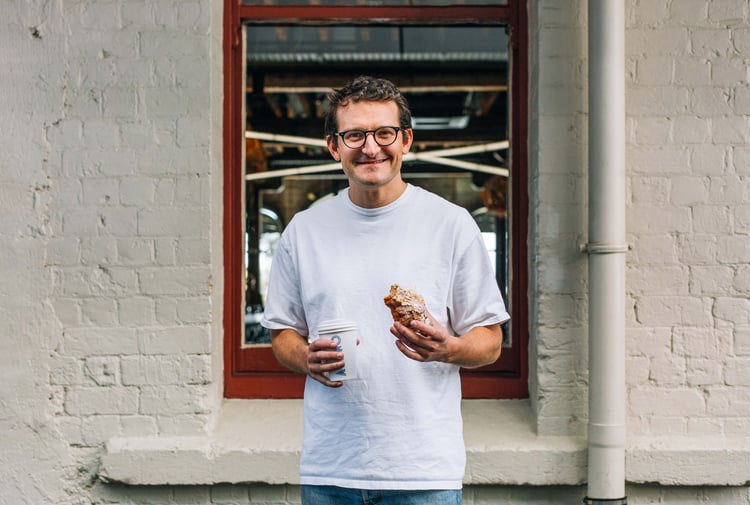 Ben Lenart holding a pastry and drink and smiling to camera.