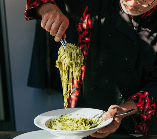 A chef serving a plate of spinach pasta on a plate in the sunlight.