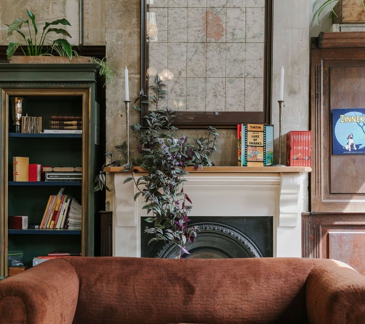 A brown suede couch in front of an old fire place decorated with books and plants.