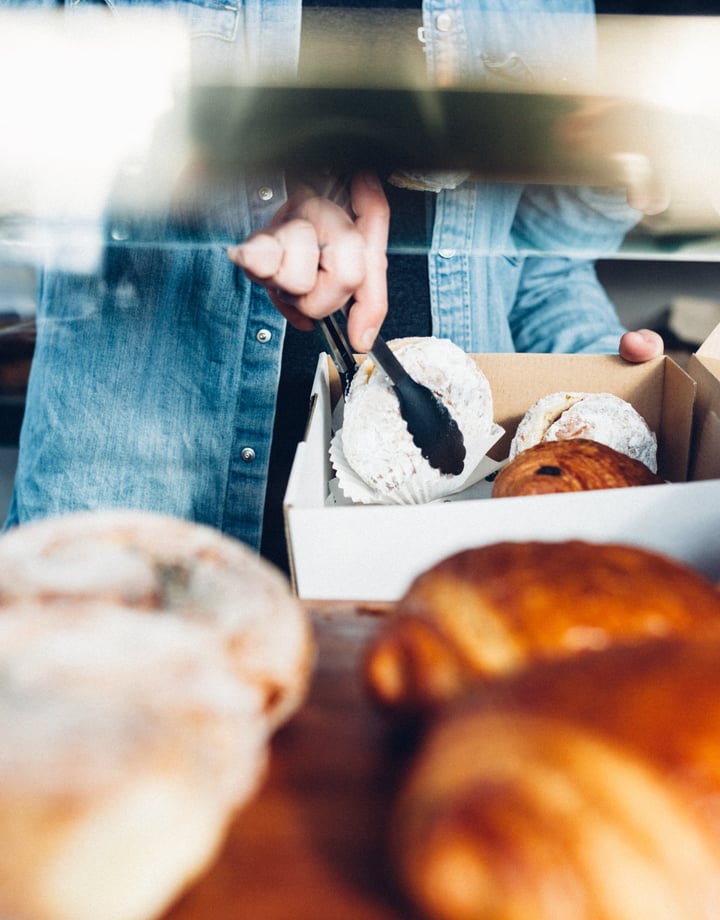 Person in denim jacket using tongs to put donuts and croissants into a take away container.