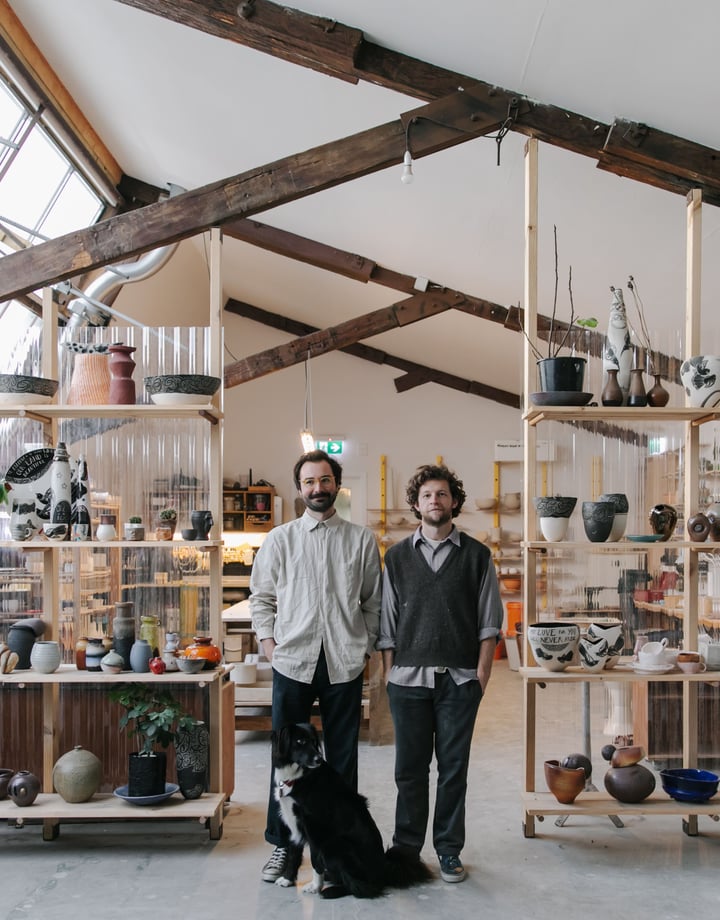 Jamie Smith and Tom Baker of Kiln Studio standing in their studio with a black dog.