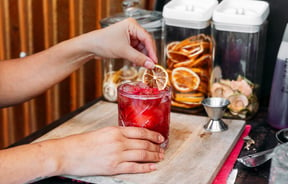 A hand holding a pink cocktail with a slice of lemon.