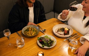 Couple enjoying plates of clam pasta, salad and steak alongside a glass of red wine