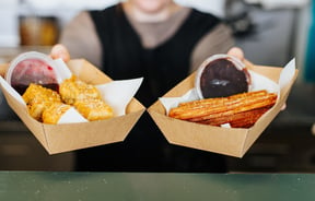 Two cardboard trays being passed over the counter; one with deep fried brownie bites and churros in the other.