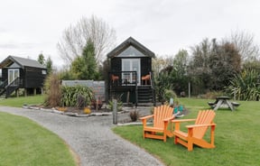 An image of a small black hut with big glass doors with deck chairs on the grass.