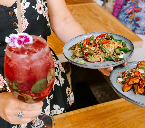 Woman holding cocktail and plates of food at Bandung Café, Auckland.