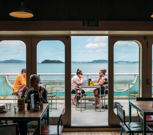 View from inside the first floor Seashore Cabaret cafe out to their deck area where people are dining with a view across the harbour to Wellington city beyond.