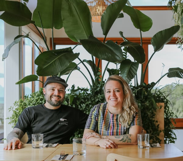 Yanina and Pablo sitting at a table under a plant, smiling to camera.