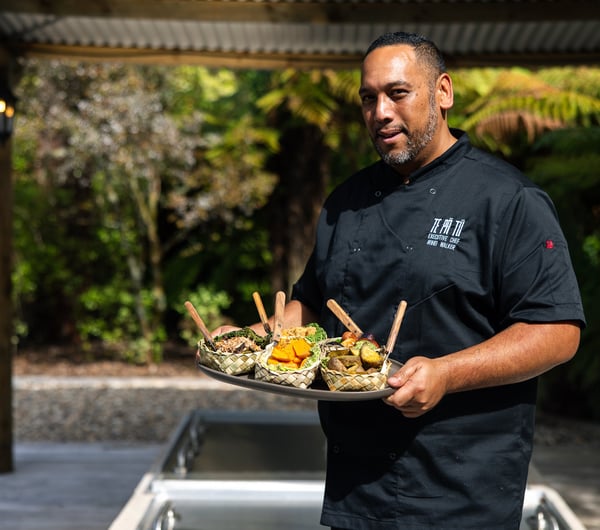 A man wearing a chef's outfit holding a tray of Maori food.