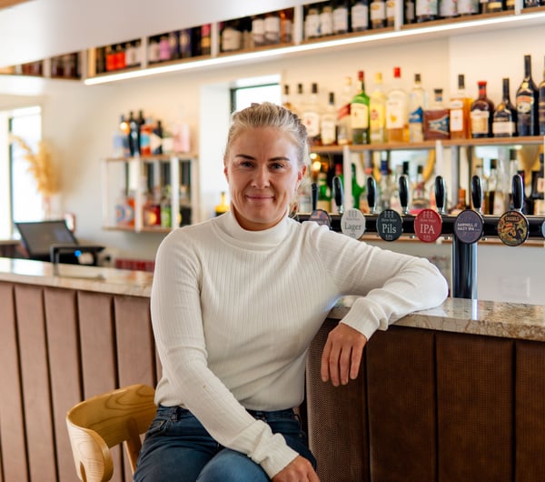 A blonde woman sitting at a bar smiling to camera.