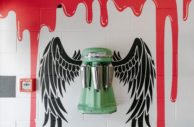 A vintage mint-green milkshake maker mounted on the wall with black wings painted behind.