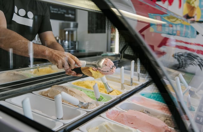 Looking into the glass gelato case where a worker is scooping gelato into a waffle cone.