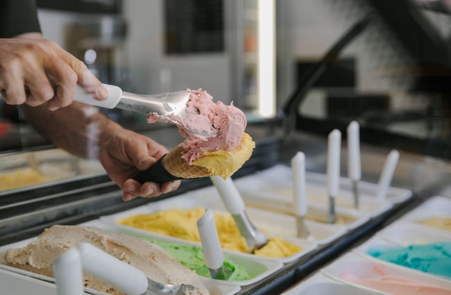 An employee scooping a pink gelato into a waffle cone with a gelato paddle.