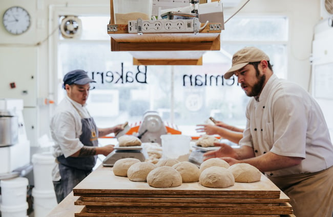 Two bakers work with bread on a large wooden bench.
