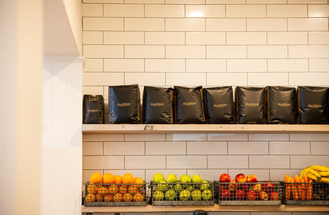 Bags of Allpress coffee on display above fresh fruit on shelves.