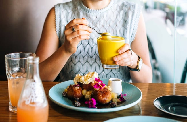 A woman sitting at a table with a turmeric latte and plate of brioche French toast.