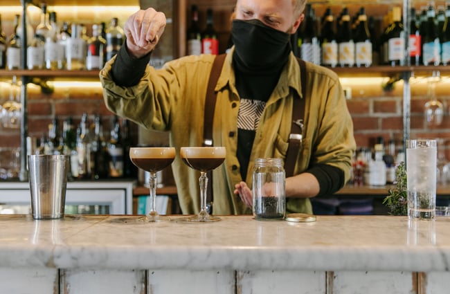 A masked staff member making two espresso martinis.