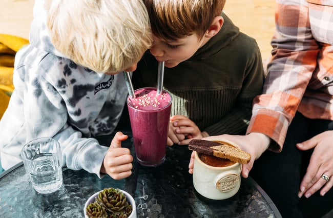 Two young boys sip a berry-coloured smoothie through metal straws and an adjacent adult holds a mug of cacao with biscotti on top.