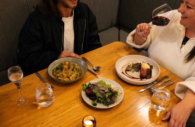 Couple enjoying plates of clam pasta, salad and steak alongside a glass of red wine