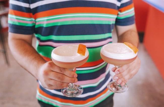 A wait staff in a bright stripe top holding two cocktails in his hands.