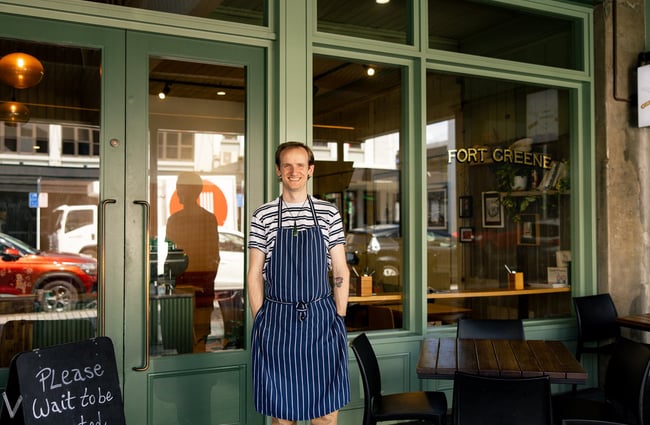 A man wearing a stripped apron smiling to camera outside his cafe.