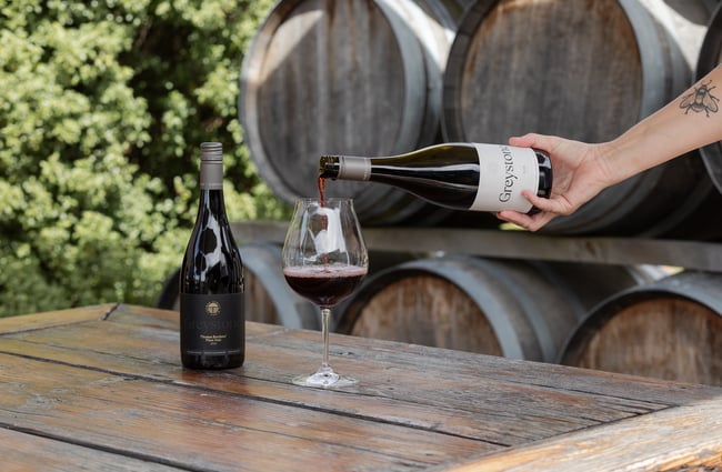 A bottle of Greystone Pinot Noir on a wooden outdoor table beside a wine glass being filled with Greystone Syrah at Greystone Wines, North Canterbury.