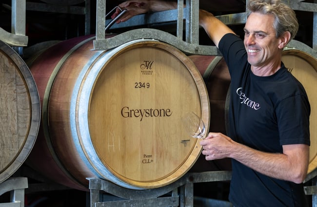 A male staff member wearing a black t-shirt with the Greystone logo on it smiles as he reaches to access a wooden wine barrel at Greystone Wines.
