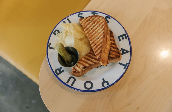 Grilled cheese sandwich with a pickle and crisps on an enamel plate at Grizzly Baked Goods in the Christchurch CBD.