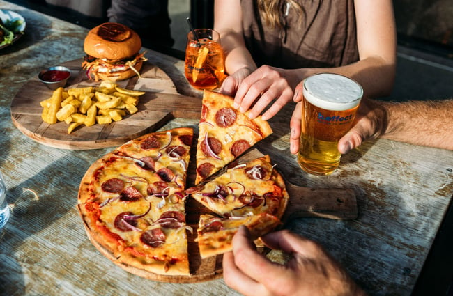 A pepperoni pizza on a wooden board on a table at hello RANGER with two people taking a slice each.