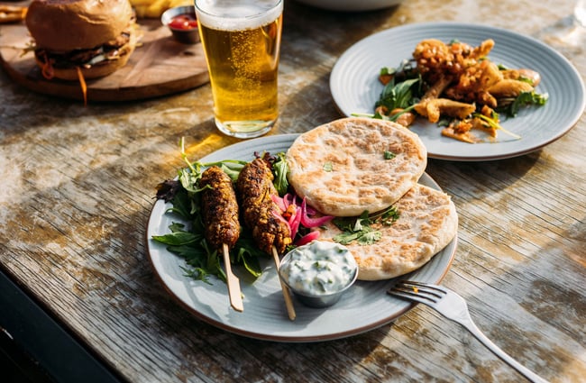 A plate of lamb koftas, flatbreads and tzatziki on a table at hello RANGER with a pint of beer and other plates in the background.