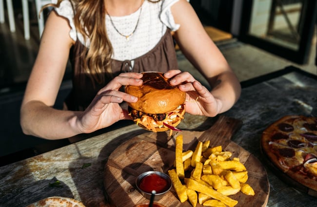 A woman holds a burger up from its wooden board with thick hot chips and tomato sauce in the sun at hello RANGER in Wānaka.